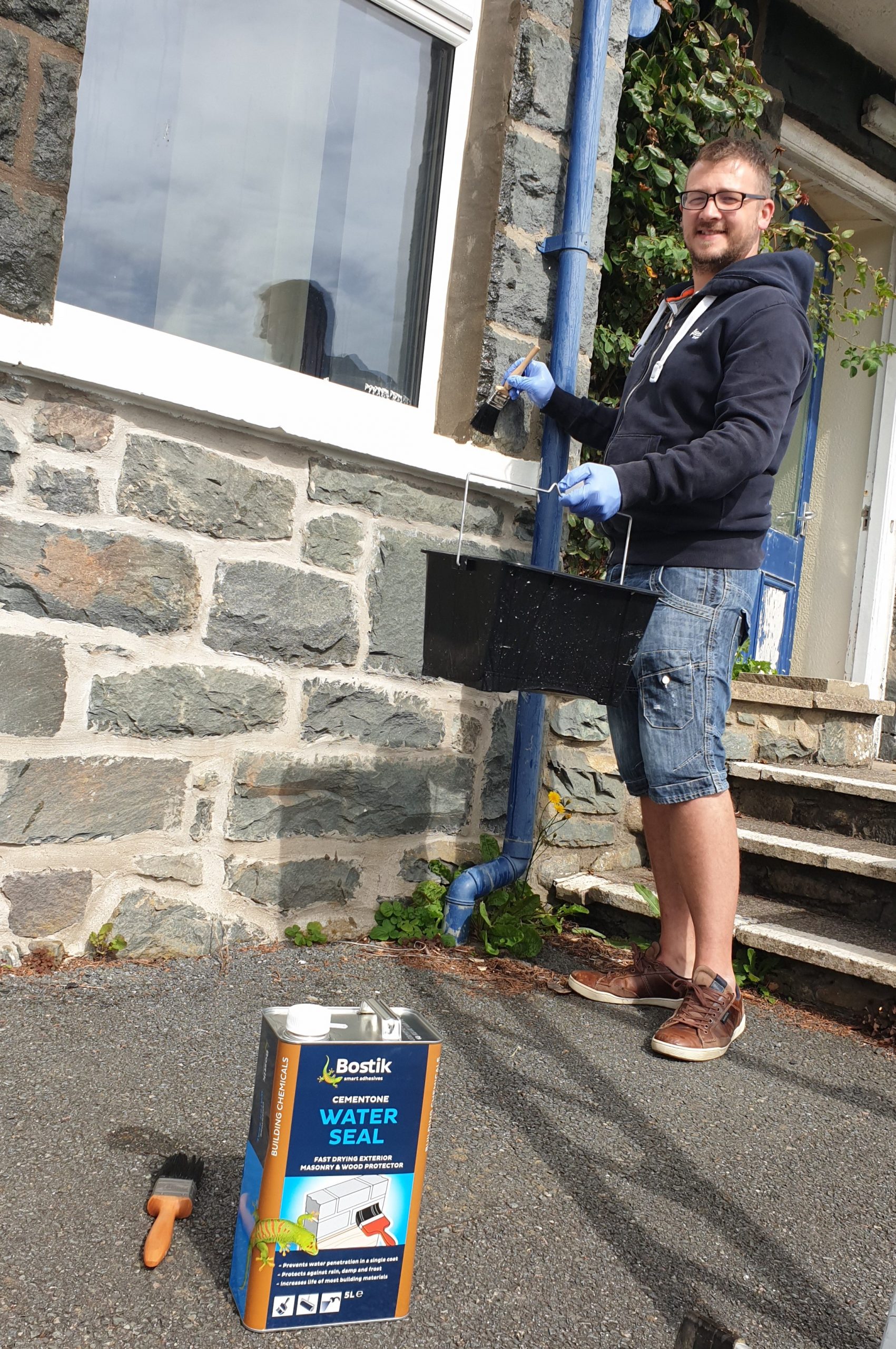 A man (me) painting a stone house with water proof clear paint. Tin in the foreground 2020